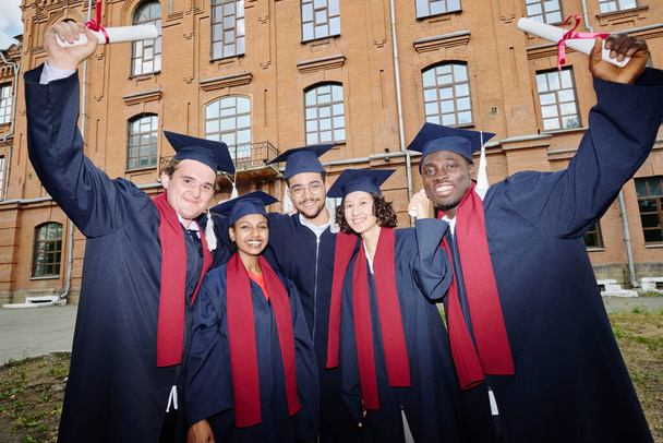 Портрет многонациональной группы выпускников в мантиях, улыбающихся в камеру, стоя на улице в кампусе - Фото, изображение
