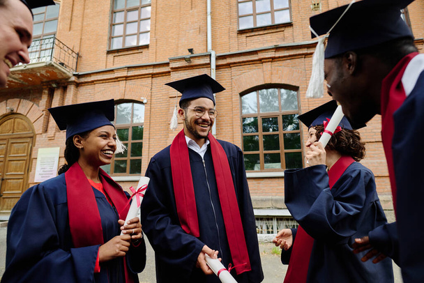 Grupa absolwentów w szlafrokach zadowolonych z uzyskania dyplomów, stojących na świeżym powietrzu i rozmawiających ze sobą - Zdjęcie, obraz