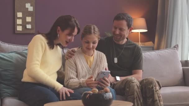 Mediana toma de alegre chica caucásica mostrando nuevo teléfono inteligente a los padres sentados entre ellos en el sofá en la sala de estar moderna y luminosa - Imágenes, Vídeo