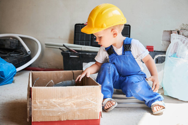 Χαριτωμένο παιδί σε κράνος ασφαλείας κάθεται στο πάτωμα κοντά σε κουτί από χαρτόνι με εργαλεία και υλικά για εργασίες ανακαίνισης και επισκευής του σπιτιού. Παιδί οικοδόμος κοιτάζοντας προμήθειες σε χαρτόκουτο. - Φωτογραφία, εικόνα