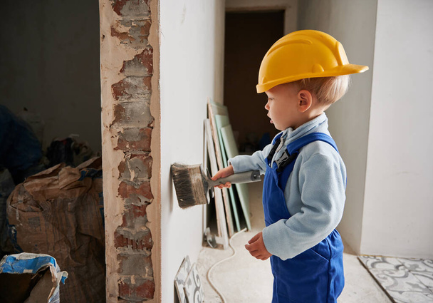 Widok z boku małego chłopca budowlańca za pomocą szczotki podczas pracy nad remontem domu. Dziecko w kasku ochronnym i kombinezonie roboczym podczas przygotowywania ściany do prac remontowych w mieszkaniu. - Zdjęcie, obraz