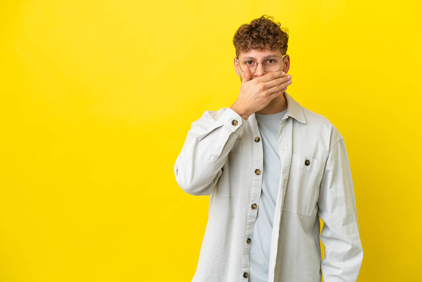 Young handsome caucasian man isolated on yellow background covering mouth with hand - Photo, Image