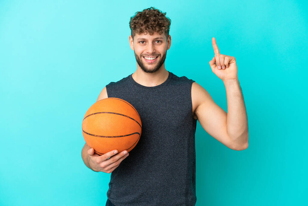 Mavi arka planda izole edilmiş basketbol oynayan yakışıklı genç adam harika bir fikre işaret ediyor. - Fotoğraf, Görsel