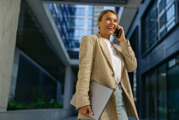Lächelnde Geschäftsfrau mit Laptop und sprechendem Telefon vor modernem Bauhintergrund  - Foto, Bild