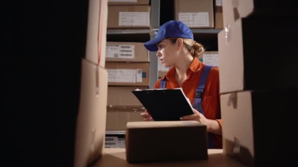 Vrouwelijke inventaris manager controleert voorraad en schrijven in het klembord, zoeken perceel. Een jonge vrouw die in een magazijn werkt. Kopieerruimte - Video