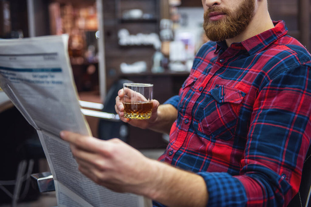 Un bell'uomo barbuto legge un giornale e tiene un bicchiere di bevanda mentre è seduto sulla sedia del barbiere.
 - Foto, immagini