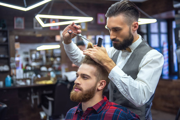 Handsome bearded man is looking forward while having his hair cut by hairdresser at the barbershop - Photo, Image
