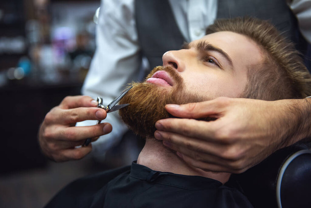 Vista laterale del bell'uomo barbuto che si fa tagliare la barba dal parrucchiere dal barbiere
 - Foto, immagini