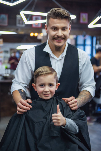 Cute little boy is showing Ok sign and smiling after having his haircut done by hairdresser at the barbershop - Photo, Image