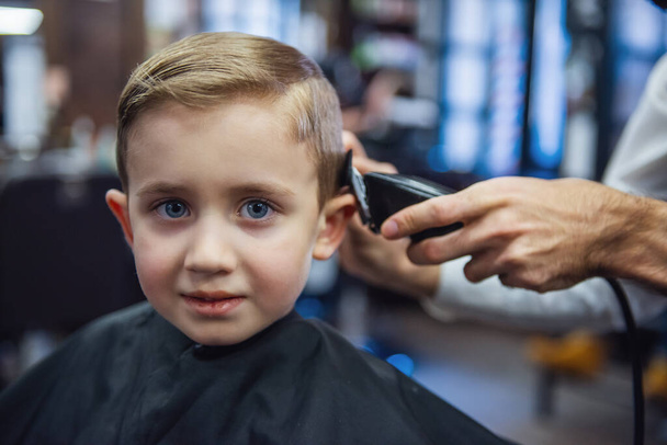Schattige kleine jongen kijkt camera terwijl ze kapsel door Kapper in de barbershop - Foto, afbeelding