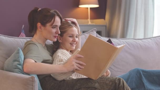 Young Caucasian soldier woman showing photo album to 11 year old daughter sitting together on couch in bright cozy living room - Footage, Video