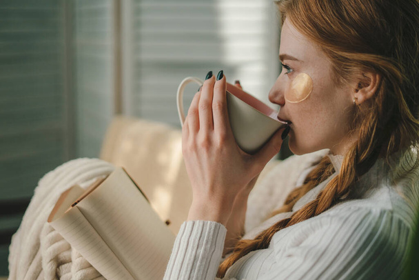 Ginger woman wrapped in woolen blanket, reading book, drinking cup of hot drink. Mature woman enjoying leisure on couch, morning coffee, studying at home. - Photo, Image
