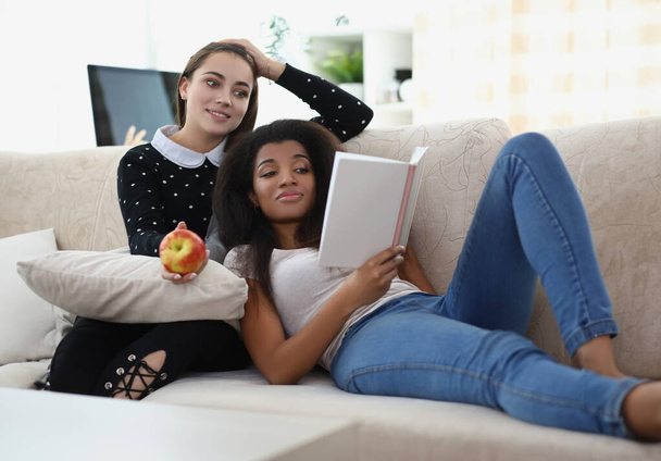 Portrait of best friends reading book chilling on sofa, apple for snack, relaxing atmosphere. Expand knowledge in literature. Leisure, friendship concept - Photo, image