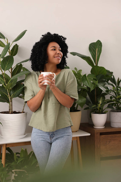 Relaxing atmosphere. Happy woman with cup of hot drink near beautiful houseplants in room - Foto, afbeelding