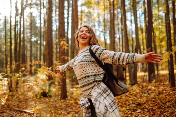 Tourist with a hiking backpack, hat walks along a path in the autumn forest. Beautiful woman enjoys a sunny day in nature, feels freedom and breathes fresh air, explores nature. - Valokuva, kuva