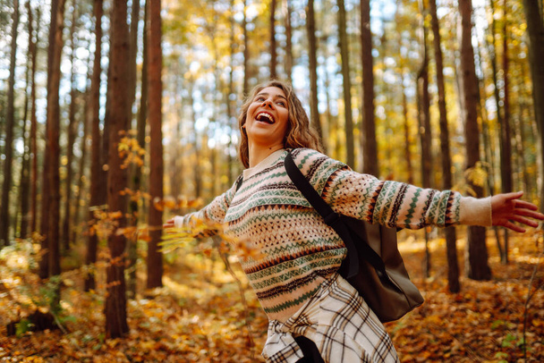 Tourist with a hiking backpack, hat walks along a path in the autumn forest. Beautiful woman enjoys a sunny day in nature, feels freedom and breathes fresh air, explores nature. - Photo, Image