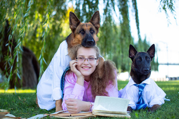 Berger Allemand et Belge (Malinois) en chemises blanches et cravates avec une fille en lunettes et une chemise rose, assis sur la pelouse verte et lisant des livres dans le parc - Photo, image
