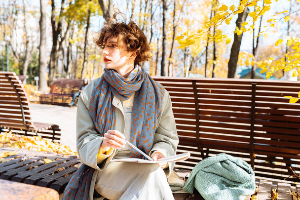 Mujer joven atractiva, con bufanda, sentado en el banco del parque, sujetando el bloc de notas para notas, sonriendo, feliz - Foto, imagen