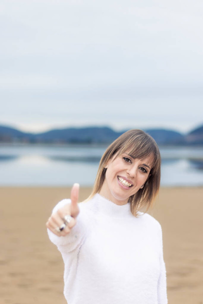 Ragyogó szépség a strandon: mosolygós nő fehér pulóverben feldobja a hüvelykujját - Fotó, kép