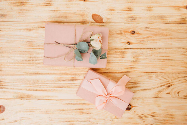 Achtergrond voor de groeten. Cadeaus verpakt in zacht roze papier met droge eucalyptustak op een lichte houten ondergrond. Bovenaanzicht - Foto, afbeelding