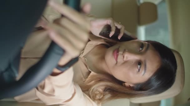 Függőleges lövés vidám ázsiai üzletasszony ül a vezetőülésen az autóban, és mosolyog, miközben beszél telefonon - Felvétel, videó