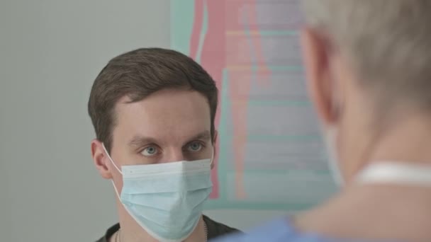 Over de schouder schot van jonge blanke man in het gezicht masker praten met arts tijdens controle afspraak in kliniek - Video