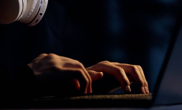 Обрезанный вид молодого компьютерного геймера в очках с наушниками на шее, печатающего на клавиатуре ноутбука, играющего в компьютерные игры, эскортирующего в темной комнате. Студент учится или общается в социальных сетях на компьютере - Фото, изображение
