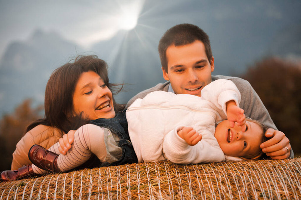 En medio de las estribaciones del campo, un niño se sienta alegremente sobre una paca de heno. Los padres cariñosos juegan y se relacionan con ella, mientras el sol de la noche los cubre con calor. - Foto, Imagen