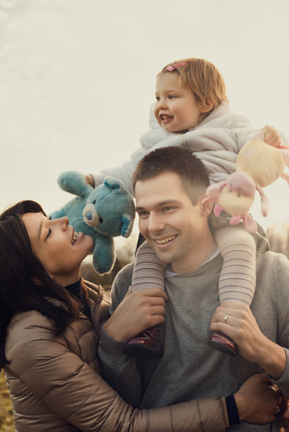 Sobre los hombros de su padre, una niña vibra de felicidad, sosteniendo sus preciados juguetes. Rodeada por el aire fresco de la caída, la madre los envuelve en el amor durante su excursión al campo - Foto, Imagen