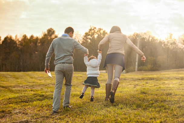 Surrounded by the golden hue of autumn, a family - man, woman, and daughter - strolls across the countryside towards a forest, enveloped in warmth, love, and divine favor - Photo, Image