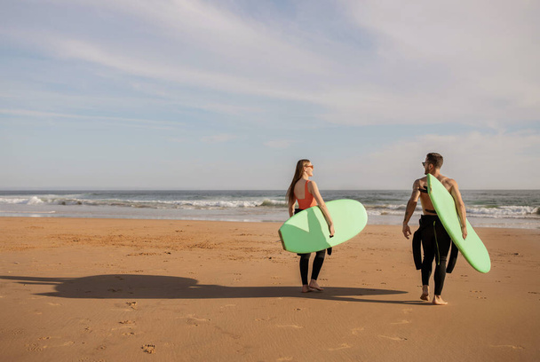Junges Surferpaar, das mit Surfbrettern an der Küste spaziert, glückliche junge Millennials in Neoprenanzügen, die ins Meer gehen, bereit zum gemeinsamen Surfen, Wassersport genießen, Raum kopieren - Foto, Bild