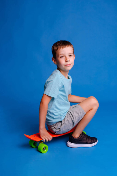 мальчик в голубой футболке сидит на скейтборде изолированный на светло-голубом фоне студии - Фото, изображение