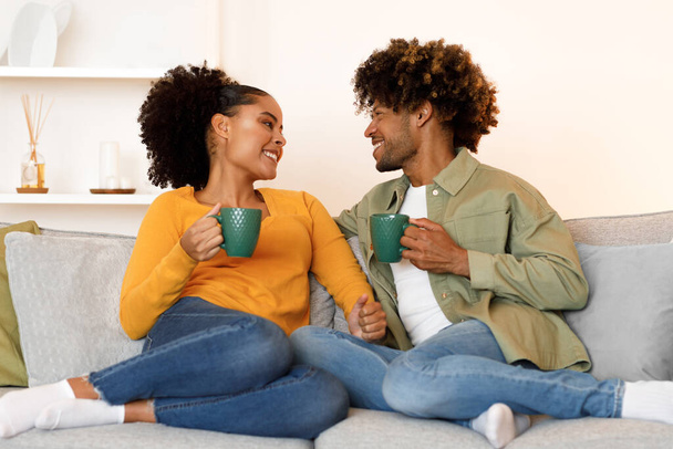 Утро выходных. Весёлая чёрная молодая пара обнимается, наслаждаясь горячим кофе на диване в помещении, держа чашки с напитками и улыбаясь друг другу. Романтика в отношениях и в быту - Фото, изображение