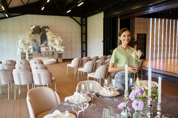 joyful florist holding vase with flowers near table with festive dishware and candles in event hall - Fotografie, Obrázek