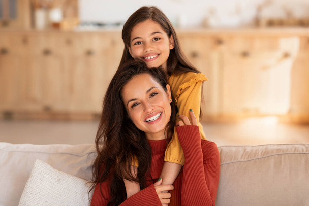 Love and motherhood. Cute middle eastern girl hugging young mommy from the back, sitting on sofa, smiling to camera, bonding at modern home interior. Portrait of kid daughter embracing her mother - Photo, Image