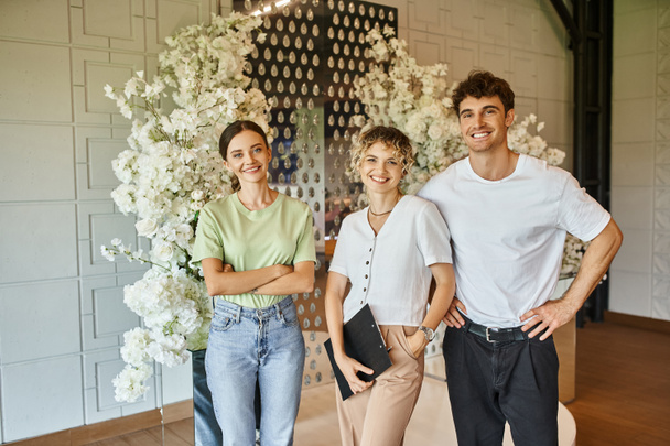 šťastný tým kreativních floristů při pohledu na kameru v hodovní síni zdobené bílými květy - Fotografie, Obrázek