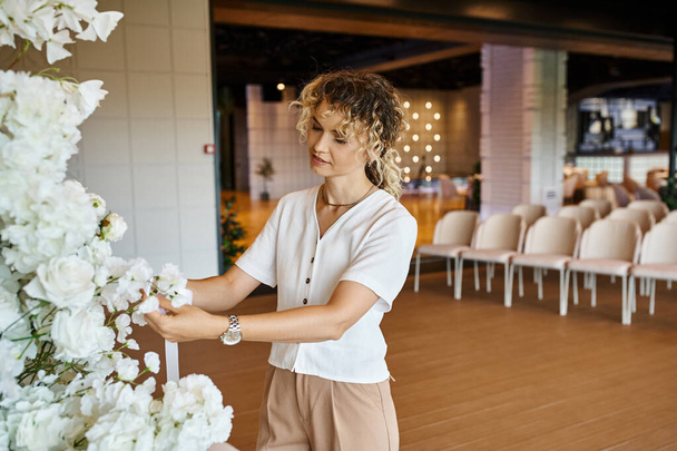 blonde florist with wavy hair arranging white floral composition in event hall, creative work - Photo, Image
