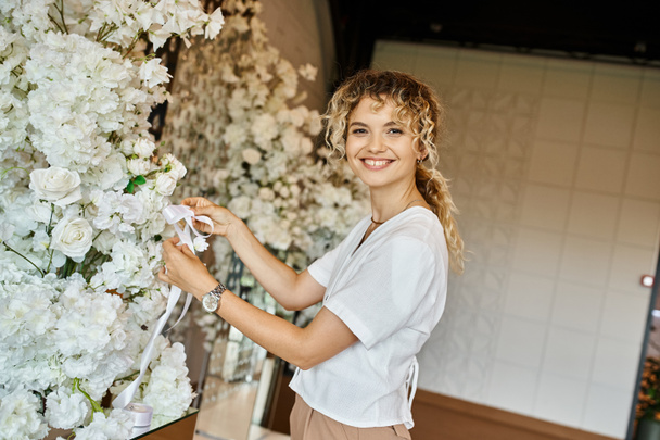 fröhliche Floristin dekoriert Veranstaltungshalle mit weißen Blumen und schaut in die Kamera, kreative Arbeit - Foto, Bild