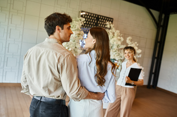 влюблённые пары смотрят друг на друга рядом с координатором мероприятия в банкетном зале с цветочным оформлением - Фото, изображение