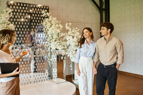 διακοσμητής δείχνει λευκό floral σύνθεση σε χαμογελαστό ζευγάρι στην αίθουσα εκδηλώσεων, προετοιμασία του γάμου - Φωτογραφία, εικόνα