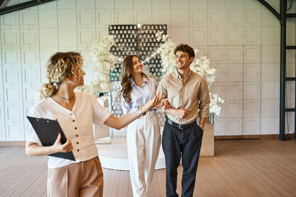 профессиональный менеджер мероприятий с буфером обмена, показывающий место проведения свадьбы с цветочным декором счастливой паре - Фото, изображение