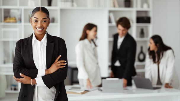 Szczęśliwa młoda, czarna bizneswoman pozująca ze złożonymi ramionami i uśmiechnięta, stojąca w sali konferencyjnej w biurze podczas spotkania z kolegami - Zdjęcie, obraz