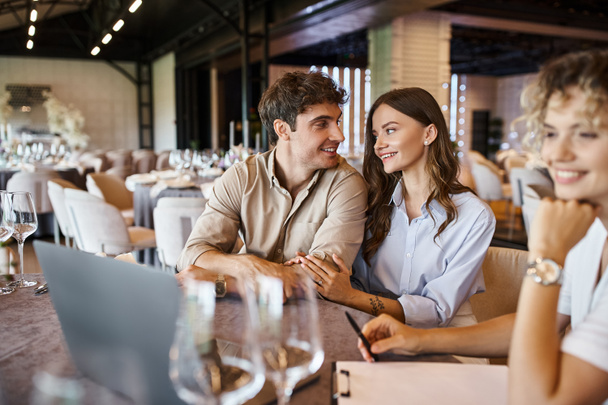 宴会場でお祝いのテーブルのラップトップの近くのぼやけたイベントマネージャーの近くで話す幸せなカップル - 写真・画像