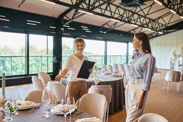 event organizer with clipboard pointing at table with festive setting near woman in banquet hall - Photo, Image