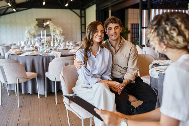 organizador do banquete com prancheta conversando com casal apaixonado no salão de eventos com decoração de casamento - Foto, Imagem