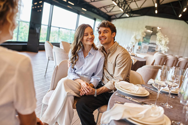 vrolijk paar in liefde zitten aan feestelijke tafel naast evenement coördinator in bruiloft locatie - Foto, afbeelding