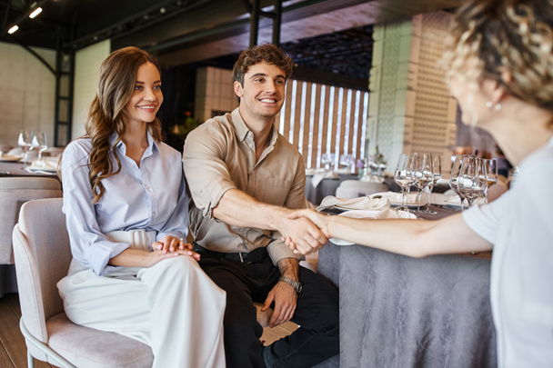 счастливый мужчина пожимает руку менеджеру мероприятия рядом с улыбающейся девушкой за праздничным столом в зале мероприятий - Фото, изображение