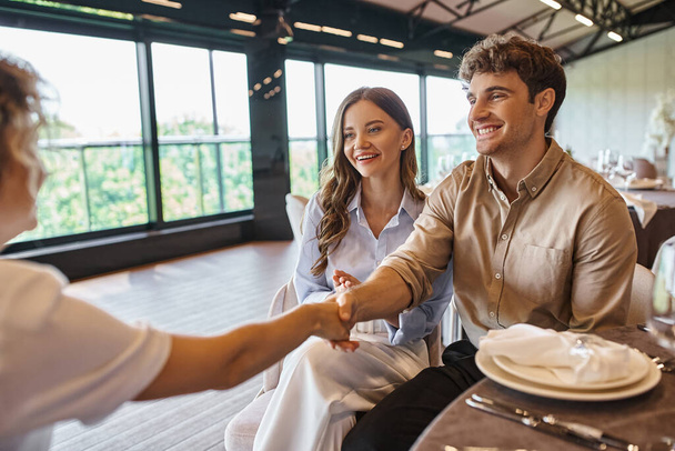 happy man shaking hands with event coordinator near pleased girlfriend in modern wedding venue - Photo, Image