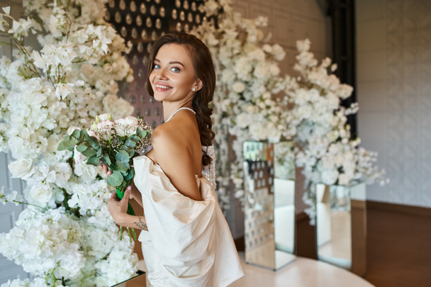 イベントホールで花の装飾の近くの花束をポーズするウェディングドレスで幸せな若い女性 - 写真・画像