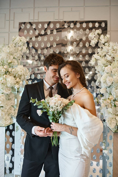 joyful couple in wedding attire with bridal bouquet in event hall decorated with white flowers - Photo, Image